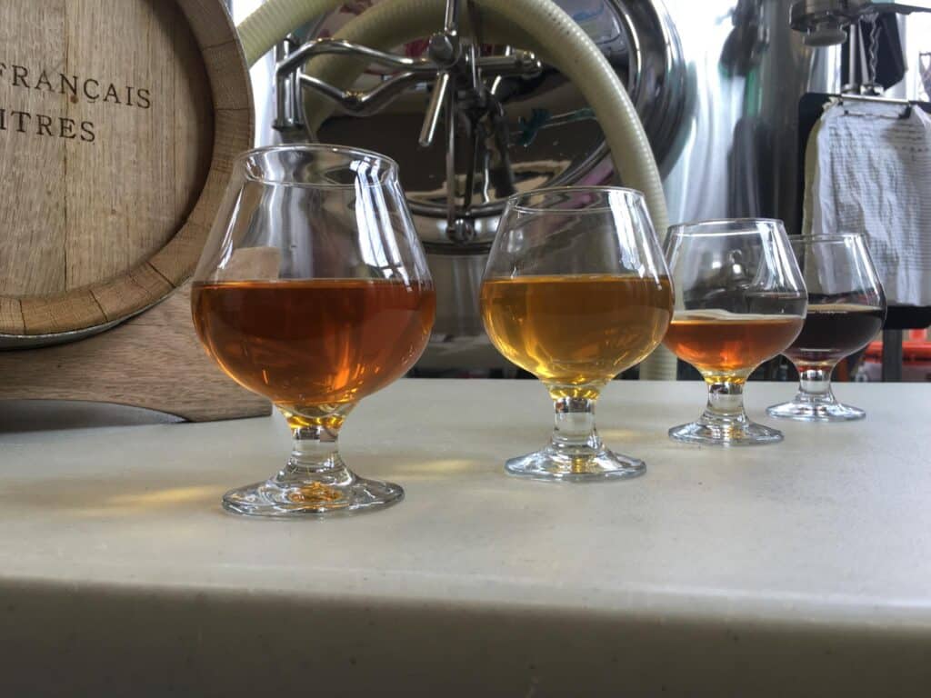 4 clear glass snifters of craft beer on white marble counter with keg and beer making equipment in background