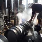 brewer leaning over silver kegs on floor of brewery with white clouds of steam rising and equipment behind