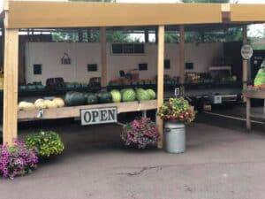 covered farm stand with white and green melons, tomatoes, flowers, open sign