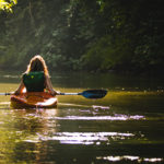 woman with long blond hair and green backpack paddling yellow kayak away on lake surrounded by green trees