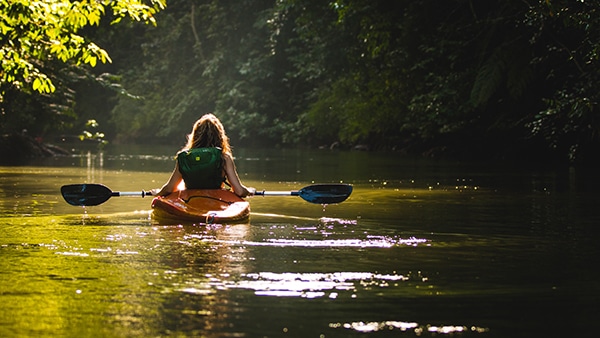 woman with long blond hair and green backpack paddling yellow kayak away on lake surrounded by green trees
