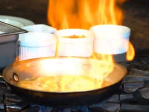 Flaming sea bass in pan with white ramekins behind