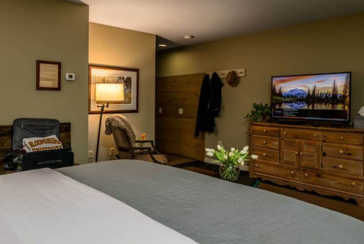 Bedroom with light taupe walls, bed with white and light sage green bedding, sitting area, and wooden dresser with large flat-screen TV