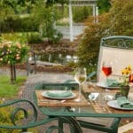 glass topped table with wine glasses and bottle & green chairs on patio in flower garden with gazebo in background