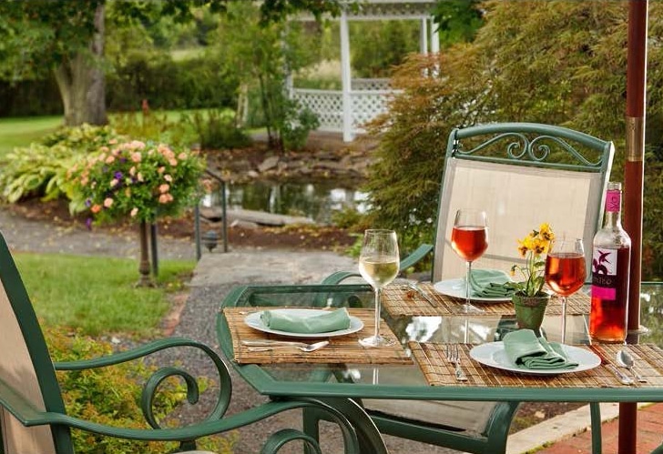 glass topped table with wine glasses and bottle & green chairs on patio in flower garden with gazebo in background