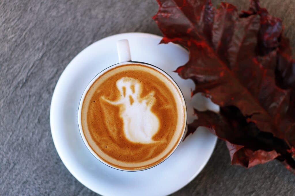 white ceramic teacup filled with ghost illustration coffee latte on white ceramic saucer beside maroon leaf