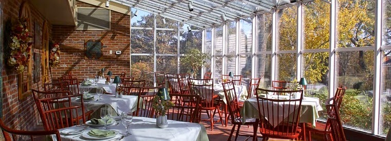 display of our restaurant green house room looking through glass windows on one side and brick on the other with tables set between for guests to arrive and enjoy fine dining