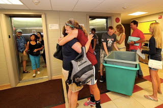 blond woman in red hugging other person in dorm hallway with room doors open and people standing around large tubs