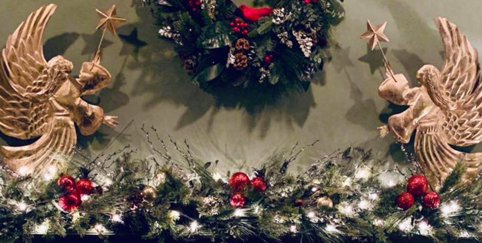 holiday decorated mantel with 2 gold angels facing central wreath, greens with white lights & red balls on mantel