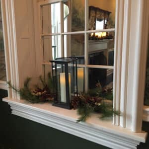 white candle in black wrought iron lantern in front of white window with christmas greens and red berries