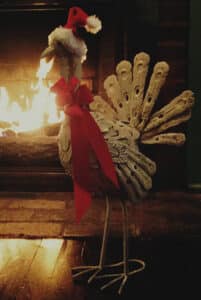 turkey sculpture standing beside blazing fireplace wearing red & white Santa hat & red ribbon around neck