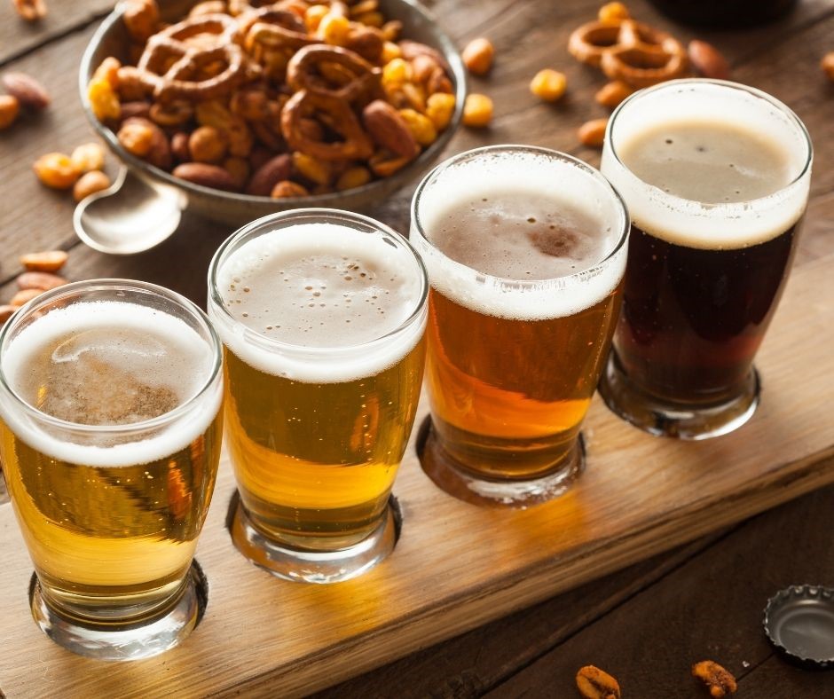 Flight of four glasses of different beer with bowl of pretzels and nuts.