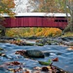 Covered bridge spanning a river with fall trees on either side.