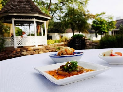 two square white plates and one white bowl with blue rim displaying food, set on white table in front of garden with white lattice gazebo, brown stone wall, green trees