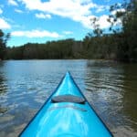 bow of turquoise kayak heading up quiet river shoreline trees & blue sky with white clouds reflected in water