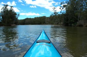 bow of turquoise kayak heading up quiet river shoreline trees & blue sky with white clouds reflected in water