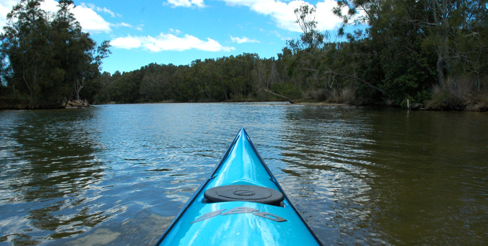 bow of turquoise kayak heading up quiet river shoreline trees & blue sky with white clouds reflected in water