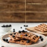 Thin stream of pouring from above on waffles with blueberries on top small white bowl of blueberries & waffle on board