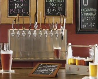 bar with glass of beer, wood framed chalkboard menu, wooden tray with beer flight on counter, 11 stainless beer taps behind on 2-toned brown wall with framed chalkboard signs above