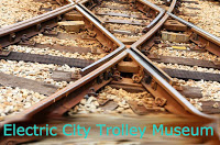 closeup of two intersecting sets of rails on brown railroad ties with lighter gravel between ties Aqua Text: Electric City Trolley Museum