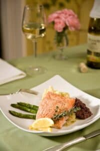 Square white plate with pink salmon filetand asparagus stalks and glass of white wine and wine bottle behind plate