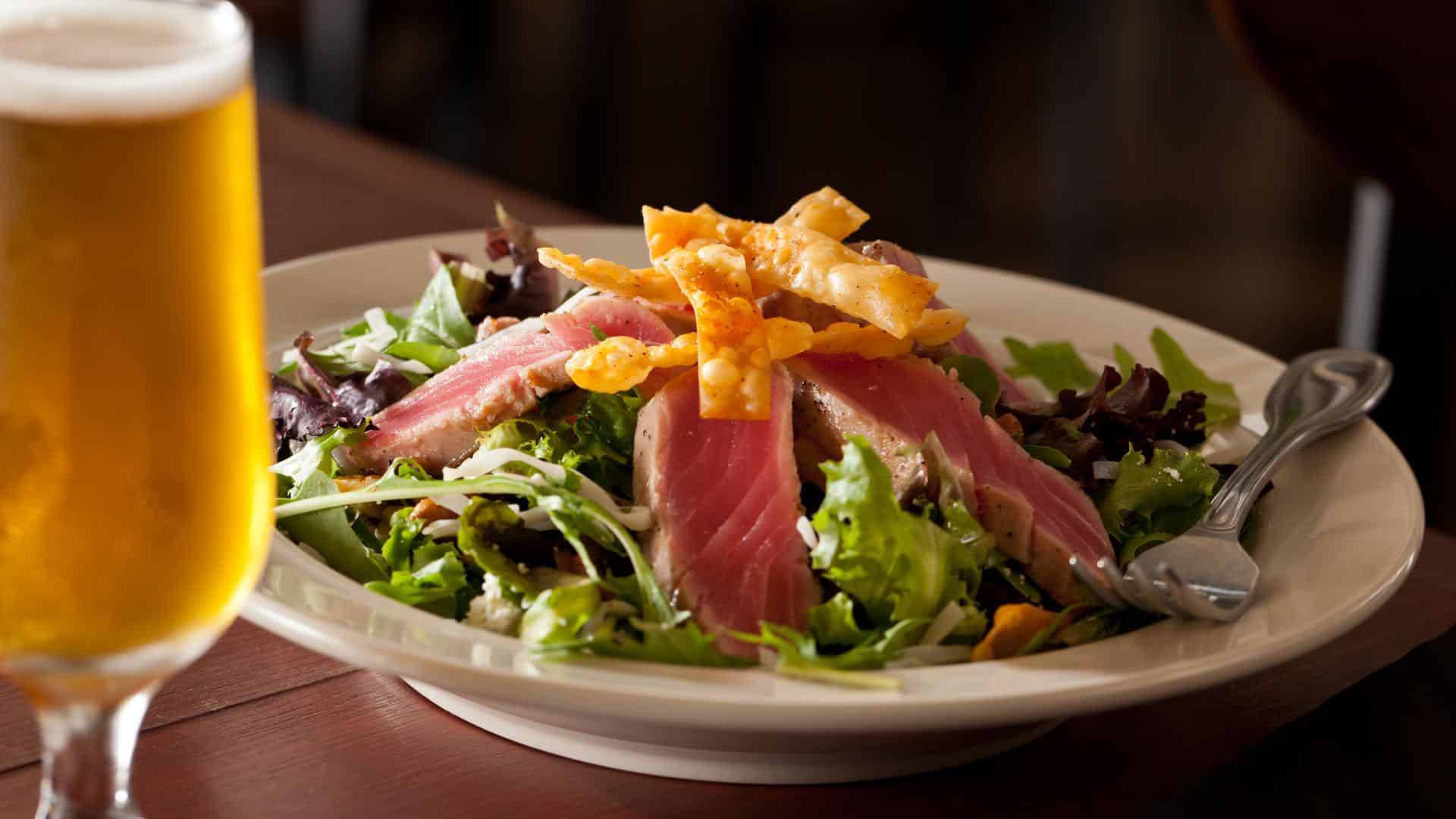 Close up view of a salad topped with grilled tuna and crispy wonton strips on a white plate next to a glass of beer