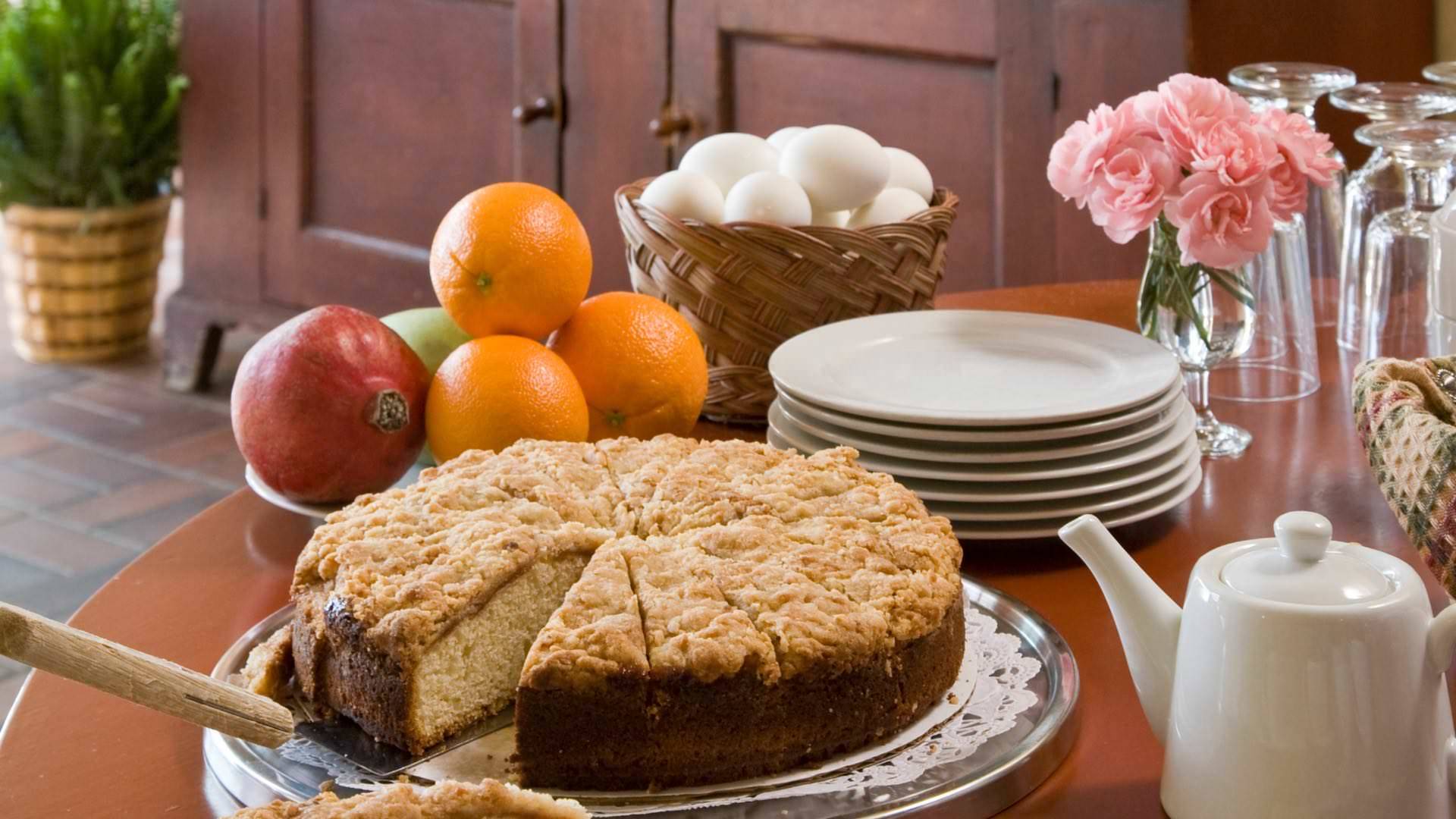 Close up view of slicked coffee cake next to a bowl of fruit, basket of eggs, small white plates, and water glasses on a round wooden table