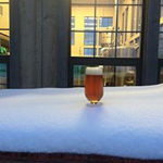 Glass of beer placed in white snowdrift on table outside lighted multi-paned window of pub at night