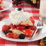 strawberry & blackberry shortcake topped with whipped cream and blueberry on white plate with fork red & white check tablecloth
