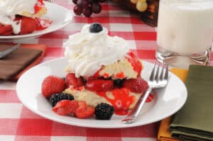 strawberry & blackberry shortcake topped with whipped cream and blueberry on white plate with fork red & white check tablecloth