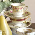 stack of three decorated fine china tea cups and saucers with gold napkin on left and white flowers in background