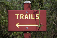 rectangular burgundy sign on post with green foliage behind Yellow Signage: Trails with arrow pointing left