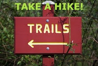 Reddish brown painted sign on post with "Trails" and arrow in yellow, green foliage behind, Green Text: Take a Hike!