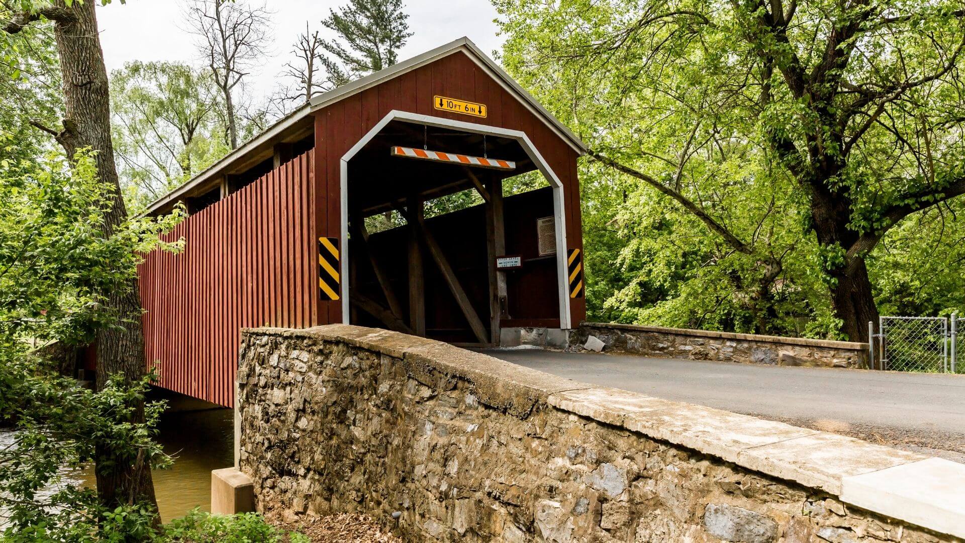 A red covered bridge crosses a creek beside stone walls and green trees.
