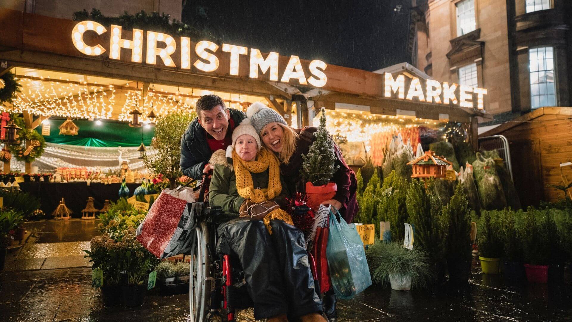 A family smiling at a Christmas market with glowing lights, shopping bags, plants, and holiday decor.