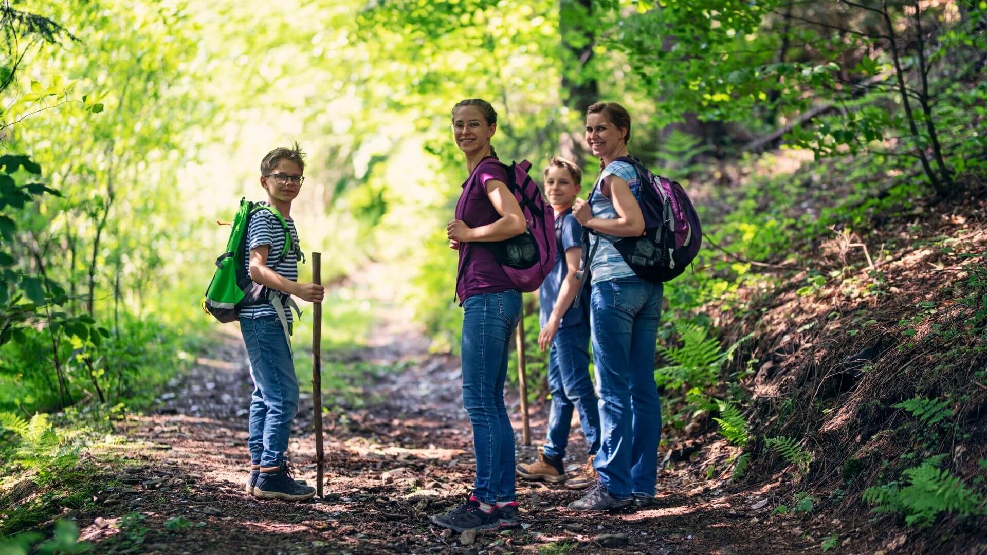 Family of four hiking on a shaded forest trail with backpacks and walking sticks, smiling and looking back toward the camera.