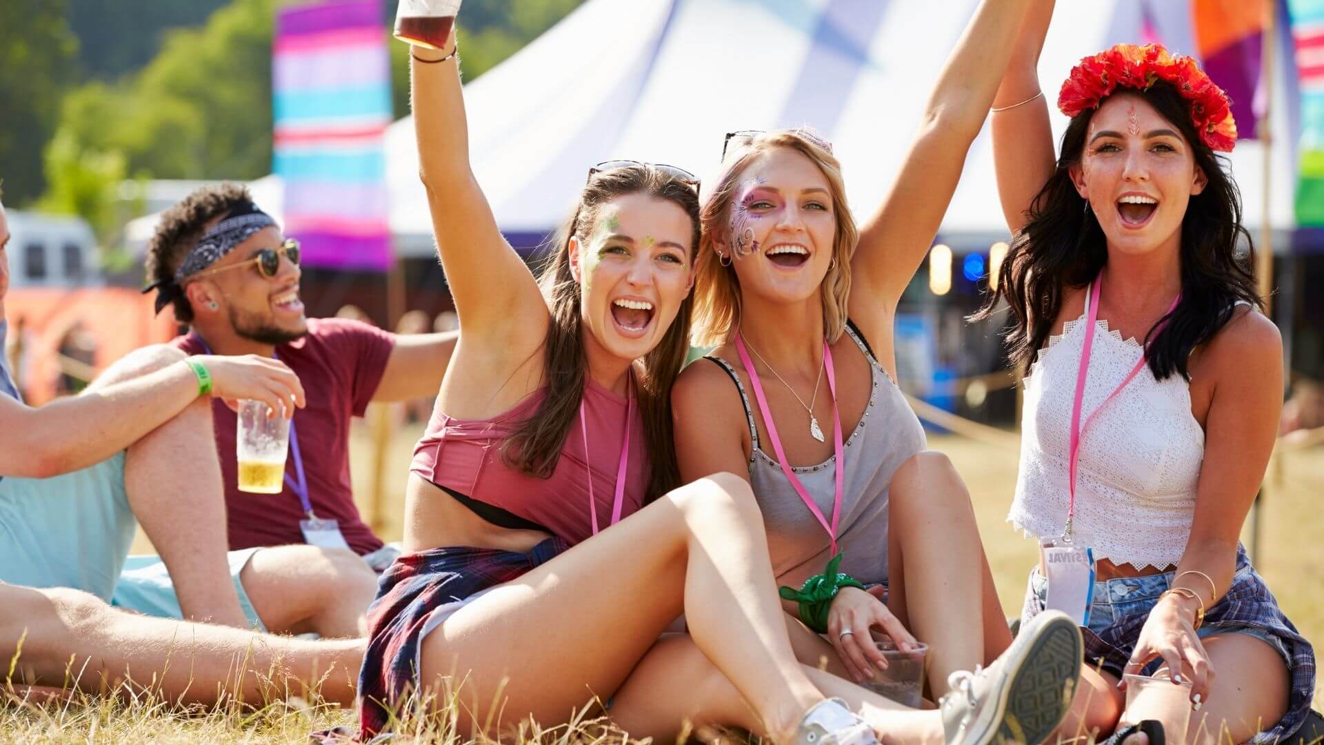 Friends sit in the grass at an outdoor festival with face paint, drinks, and colorful tents nearby.