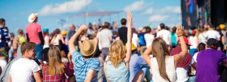 group of friends sitting on grass at an outdoor music festival with arms raised facing the stage under a bright blue sky