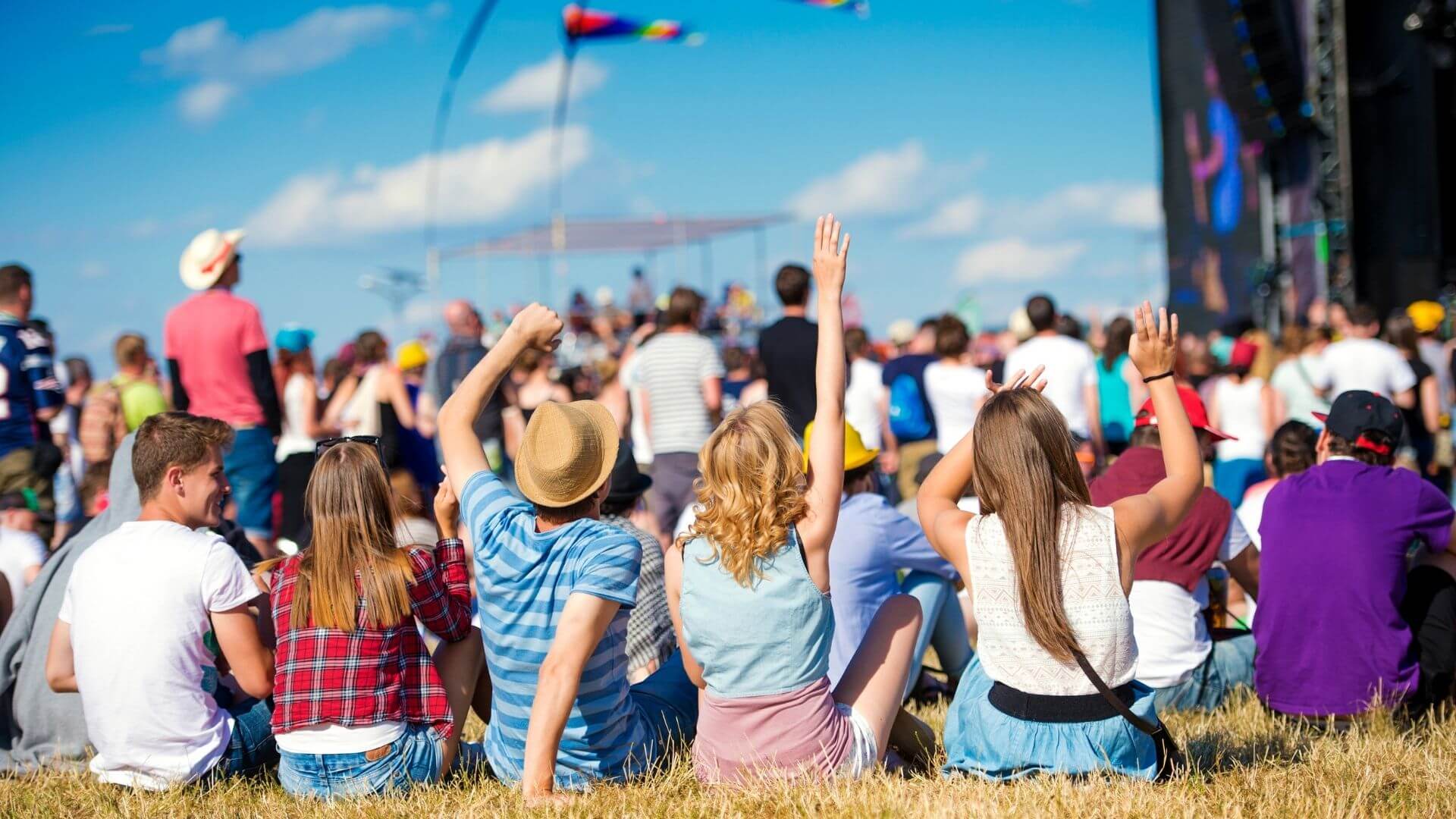 group of friends sitting on grass at an outdoor music festival with arms raised facing the stage under a bright blue sky