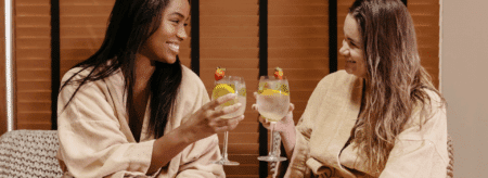 two women in spa robes smiling and toasting with cocktails garnished with strawberries