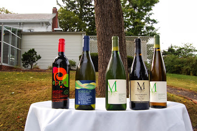 5 bottles of wine with red, blue, white, beige labels placed on circular table with white cloth, in front of tree trunk, white building with greenhouse behind
